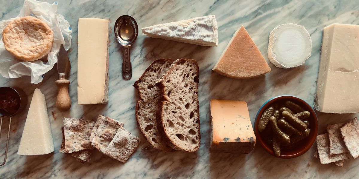 An overhead cheese board on a stone surface with blue cheese, brie, cheddar, sliced bread, oatcakes, strawberries, grapes, apricots and rosemary