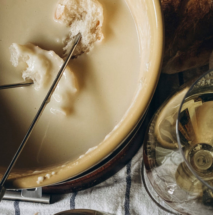 A close-up overhead view of a cheese fondue scene with a cream-colored cheese bowl, metal cheese forks, bread, and wine glasses on a striped cloth.