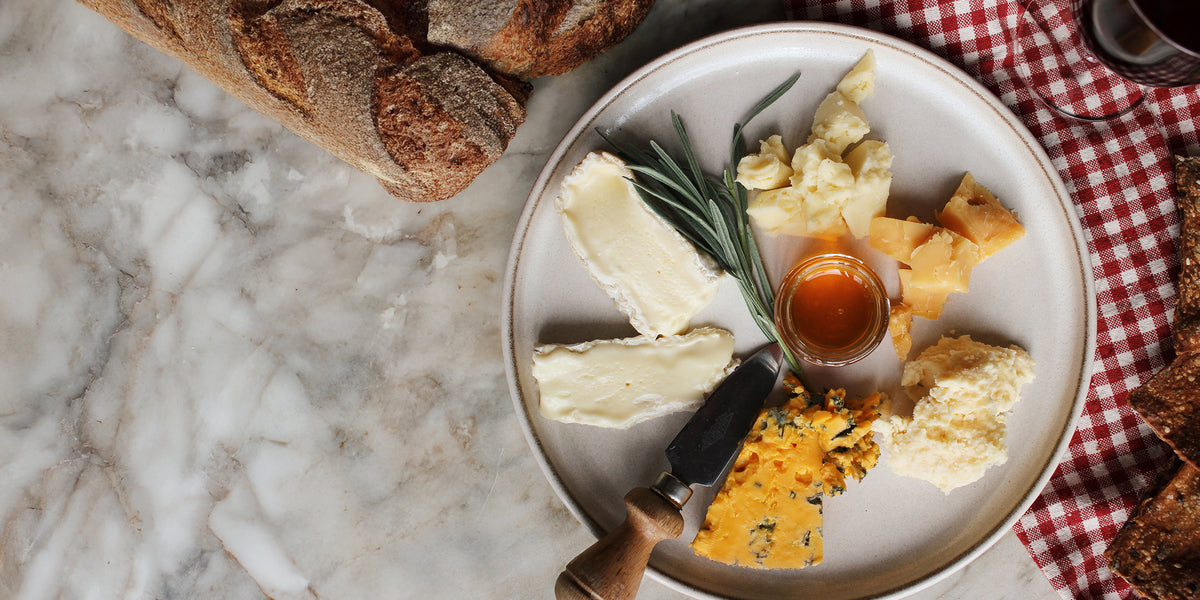 An overhead cheese board on a stone surface with blue cheese, brie, cheddar, sliced bread, oatcakes, strawberries, grapes, apricots and rosemary