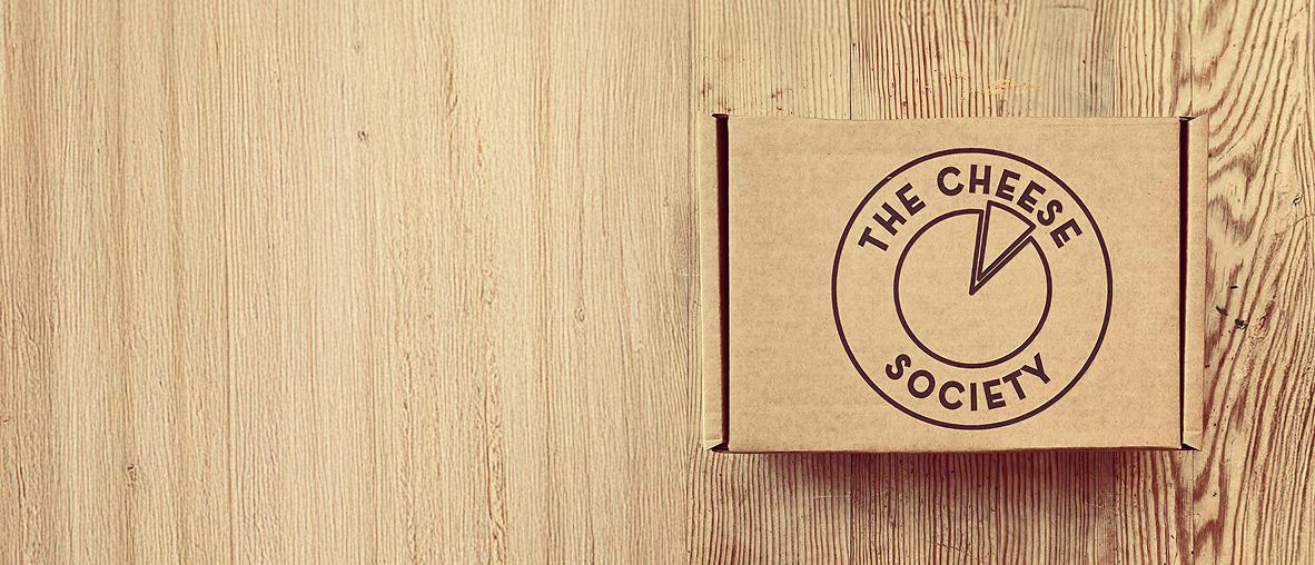 A Cheese Society cardboard delivery box with the circular logo, seen from above on a pale wooden table